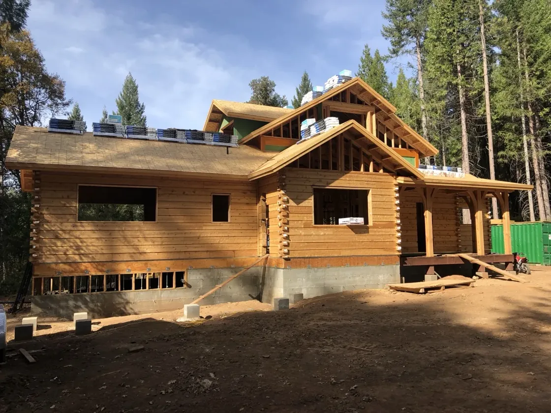 A single-storey wing meeting the taller centre of the house, log walls up and the roof decked, bundles of roofing staged along the ridge and tall pines all around.