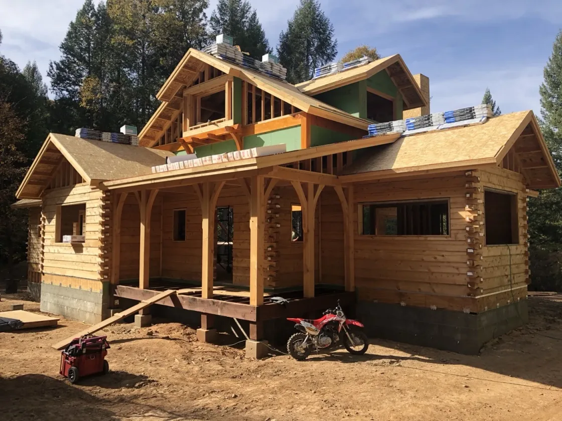 A log and timber shell nearly ready for roofing, with arched porch timbers, green-sheathed upper walls and bundles of shingles staged along the roof edges.