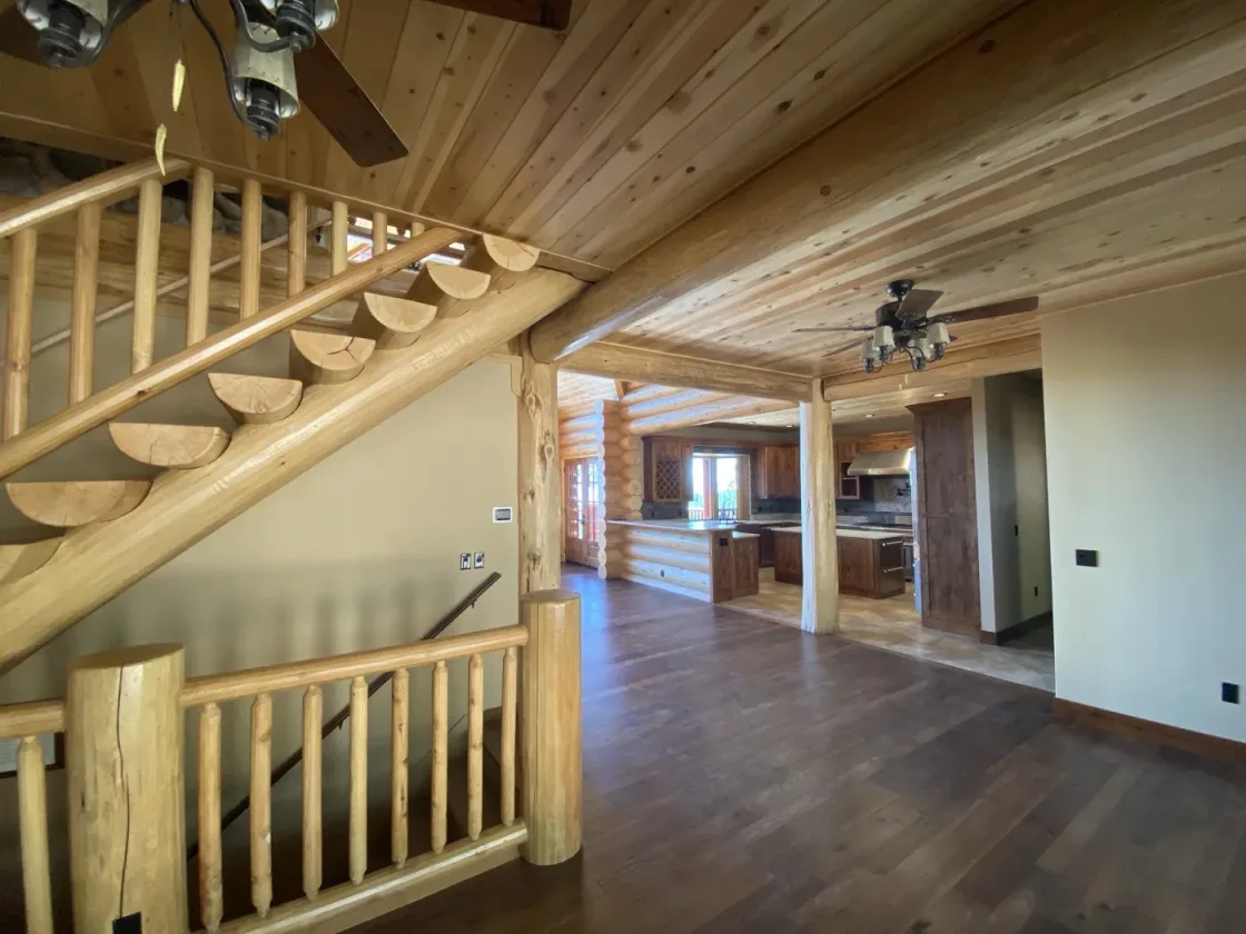 Inside a finished home: a log staircase with half-log treads, wood-plank ceilings on log beams, and the kitchen beyond the great room.