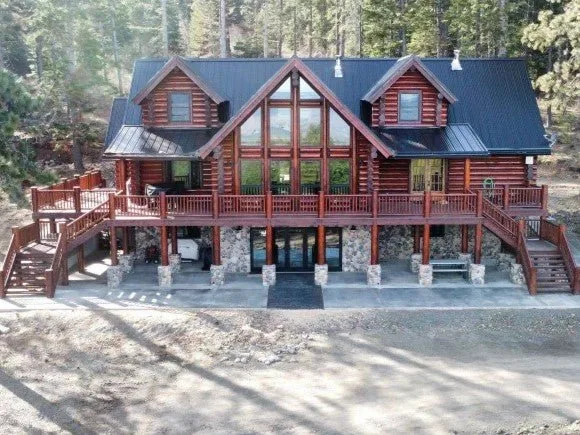A finished two-storey log home with a prow front of windows, a full-width deck on stone piers, stairs at both ends and a dark metal roof against the pines.