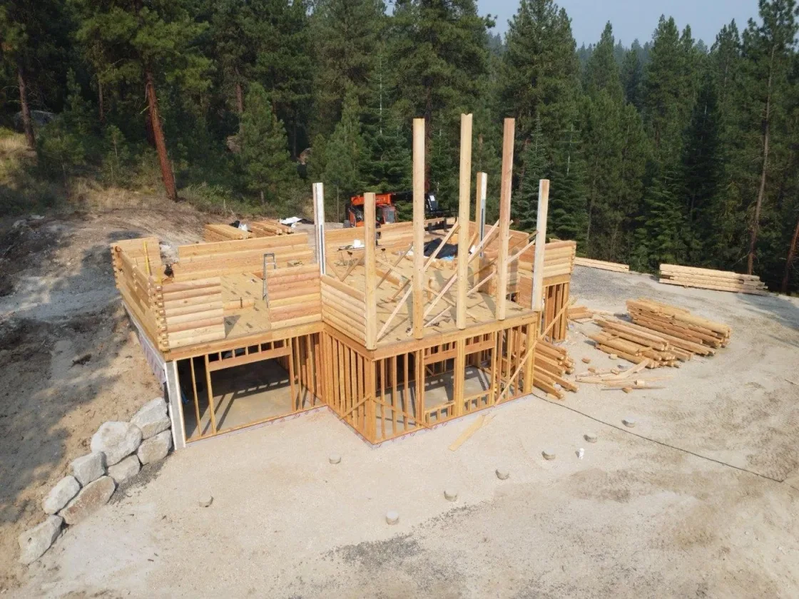 A build seen from the air: the log-walled first storey decked over, roof posts standing, and bundles of logs staged on the gravel pad among the pines.