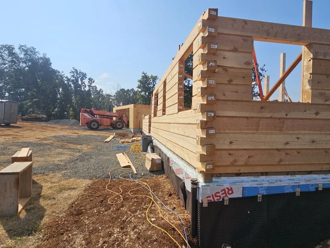The corner of a squared-log wall up close, each course end-tagged with its number, window openings framed and an orange telehandler parked across the site.